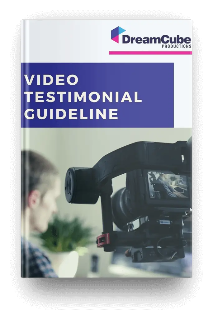 The image features a printed guide titled "VIDEO TESTIMONIAL GUIDELINE" with the DreamCube Productions logo at the top. The cover showcases a blurred photo of a professional video camera on a tripod with a person in the background, indicating the guide's focus on creating video testimonials. The guide seems to be lying against a white backdrop, suggesting a clean and simple presentation style.