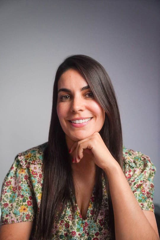 DreamCube copywriter Christelle Delli smiles, chin on hand, floral blouse, grey background.