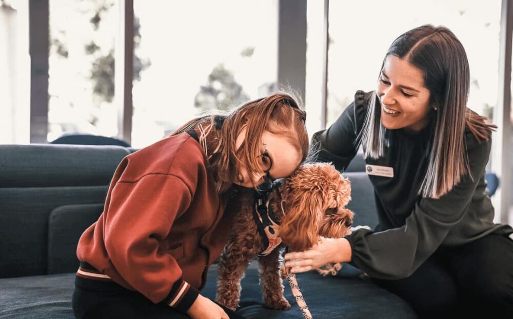 Village Therapy Corporate Video: Girl in red hoodie nuzzles fluffy dog, woman facilitates in comforting room.